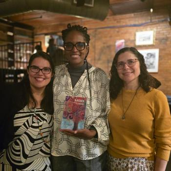 Three women standing. The middle one is holding the poetry in translation book Raizes